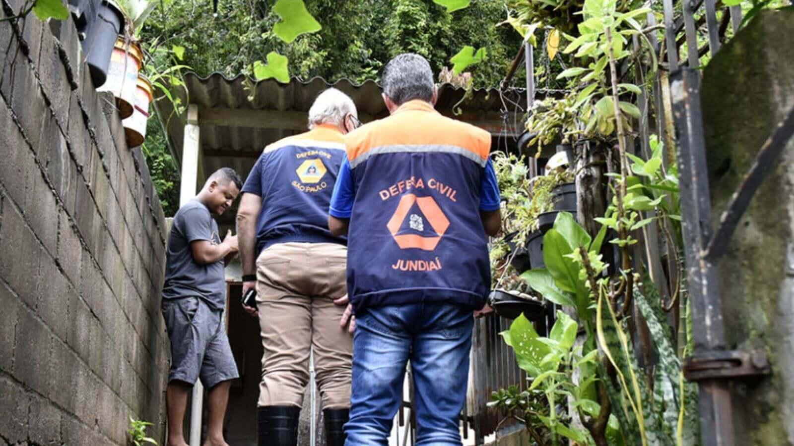 Dois homens vestidos com coletes da Defesa Civil de Jundiaí em uma área externa, possivelmente em um quintal ou um local com plantas e vegetação.