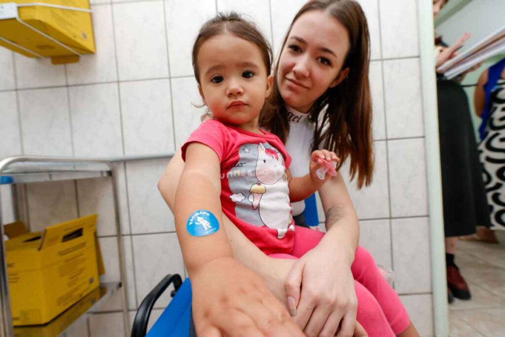 Criança mostra adesivo "vacinado" no braço enquanto é segurada por uma mulher, após receber vacina contra poliomielite em uma clínica de saúde.
