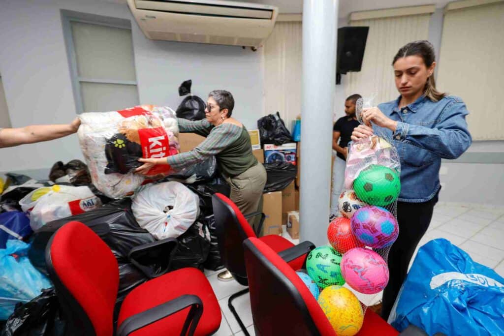 Doações de Jundiaí para crianças vítimas das enchentes no RS. Pessoas organizam brinquedos e mantimentos em uma sala cheia de sacolas e caixas.