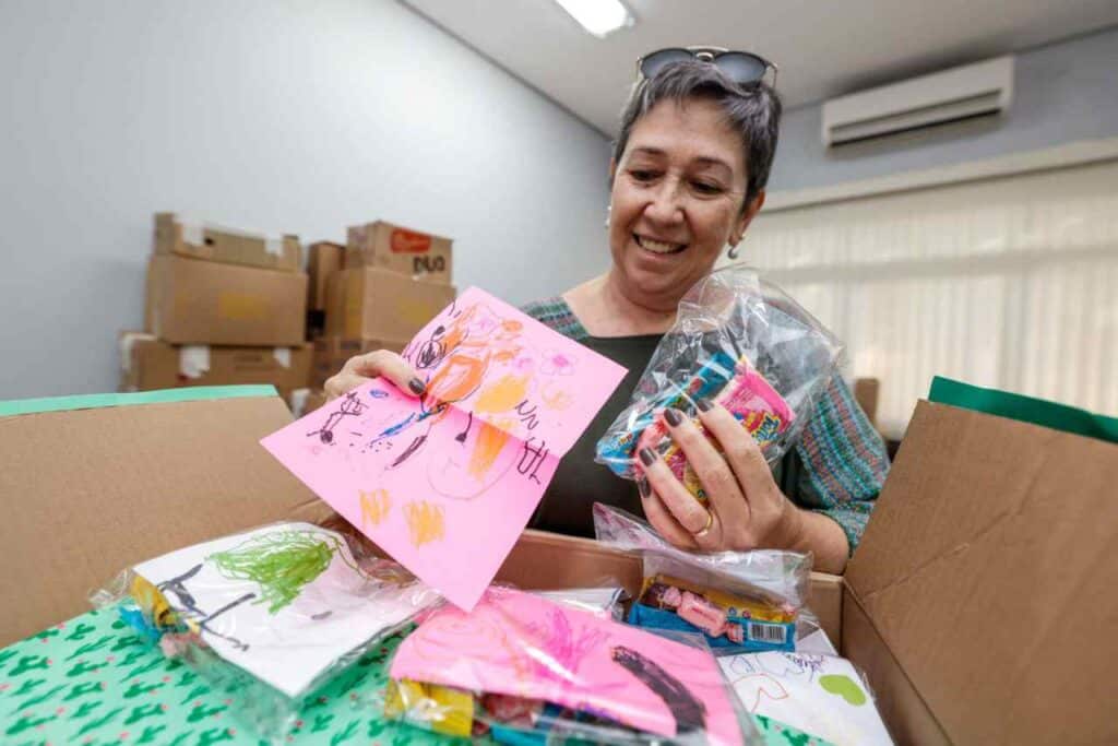 Doações de Jundiaí para crianças vítimas das enchentes no RS. Mulher sorri ao organizar desenhos infantis e brinquedos em uma caixa.