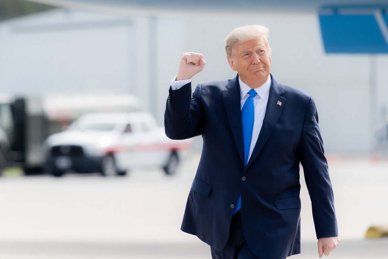 Donald Trump em um terno escuro e gravata azul levanta o punho em um gesto confiante, com um leve sorriso no rosto, parado ao ar livre perto de veículos.