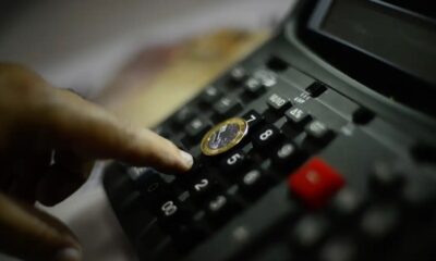 Pessoa utilizando uma calculadora com moeda de um real, representando economia brasileira