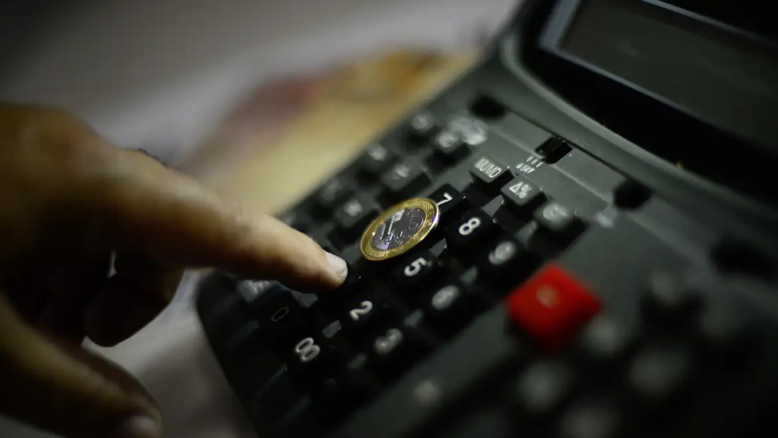 Pessoa utilizando uma calculadora com moeda de um real, representando economia brasileira