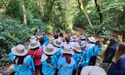 Um grupo de estudantes em uniformes azuis e chapéus bege está em uma trilha na Serra do Japi, participando de uma aula ao ar livre sobre mudanças climáticas.