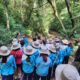 Um grupo de estudantes em uniformes azuis e chapéus bege está em uma trilha na Serra do Japi, participando de uma aula ao ar livre sobre mudanças climáticas.