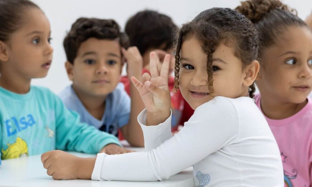 Grupo de crianças sentadas à mesa no Lar Creche Wilson de Oliveira, em Jundiaí. Em primeiro plano, uma menina de cabelo cacheado sorri para a câmera e faz o sinal de paz com os dedos.