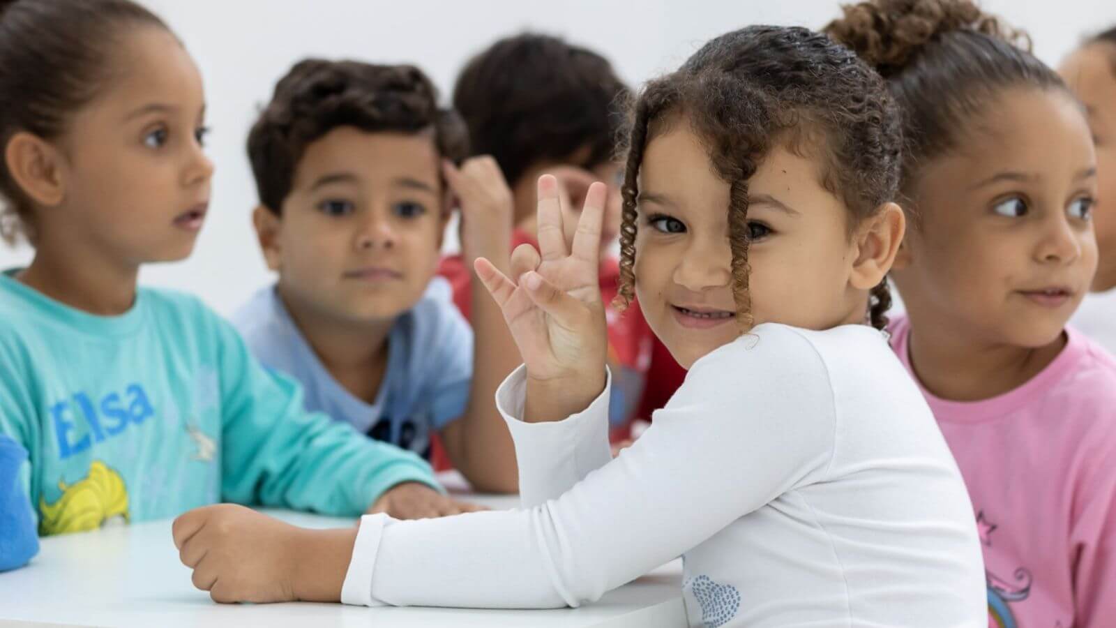 Grupo de crianças sentadas à mesa no Lar Creche Wilson de Oliveira, em Jundiaí. Em primeiro plano, uma menina de cabelo cacheado sorri para a câmera e faz o sinal de paz com os dedos.