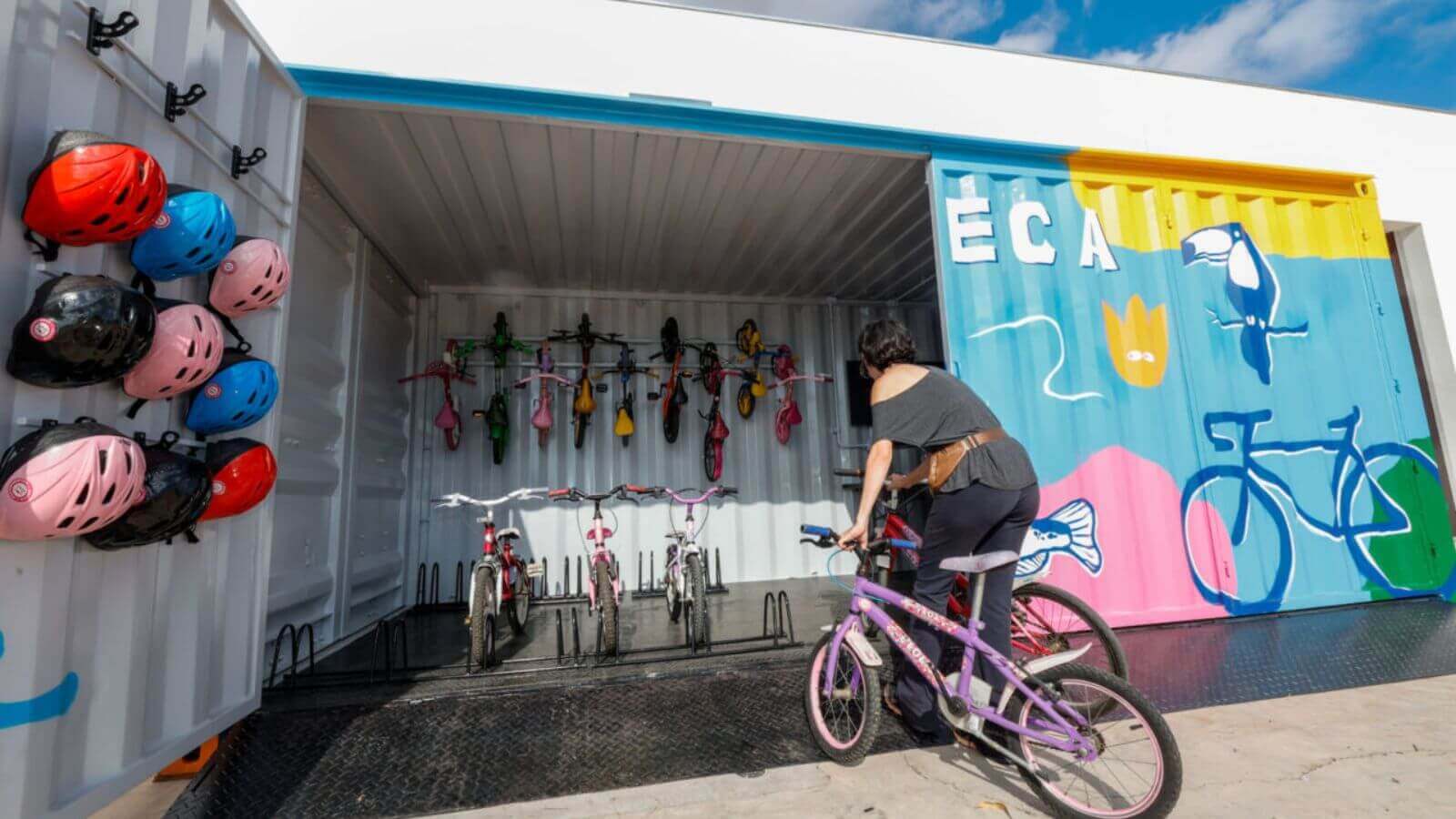 Imagem de uma mulher pegando uma bicicleta em uma bicicloteca de Jundiaí, que possui capacetes coloridos pendurados e bicicletas organizadas dentro de um contêiner decorado.