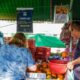 Clientes comprando frutas frescas em barraca de feirantes regularizados em Jundiaí, com cartaz destacando feirantes licenciados ao fundo.