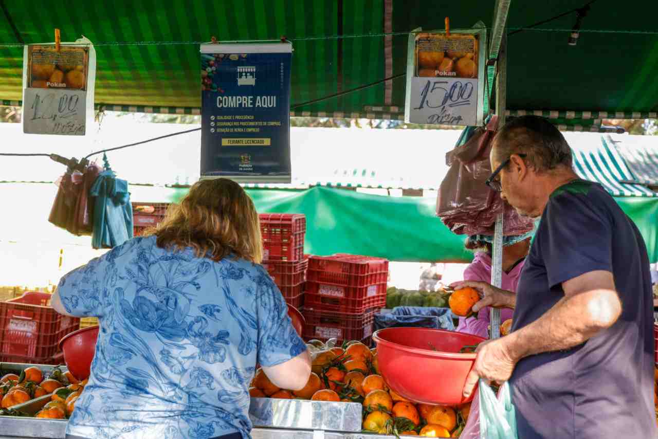 Clientes comprando frutas frescas em barraca de feirantes regularizados em Jundiaí, com cartaz destacando feirantes licenciados ao fundo.