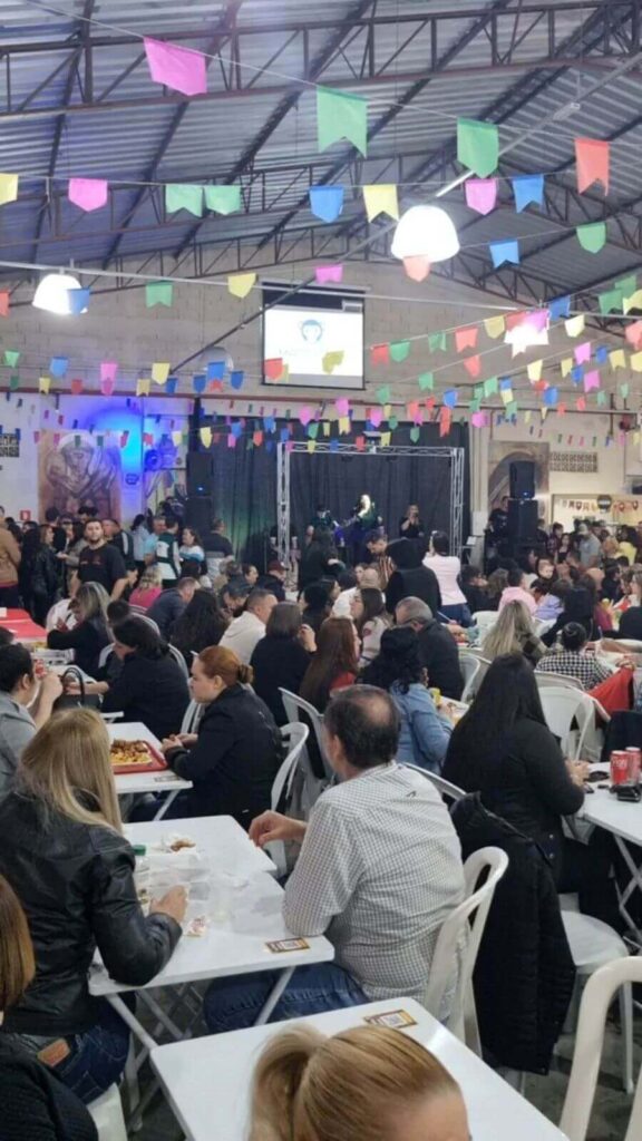 Festa decorada com bandeirinhas no teto, típicos de festas juninas ou comemorações religiosas. Muitas pessoas estão sentadas em mesas em paróquia de Jundiaí.