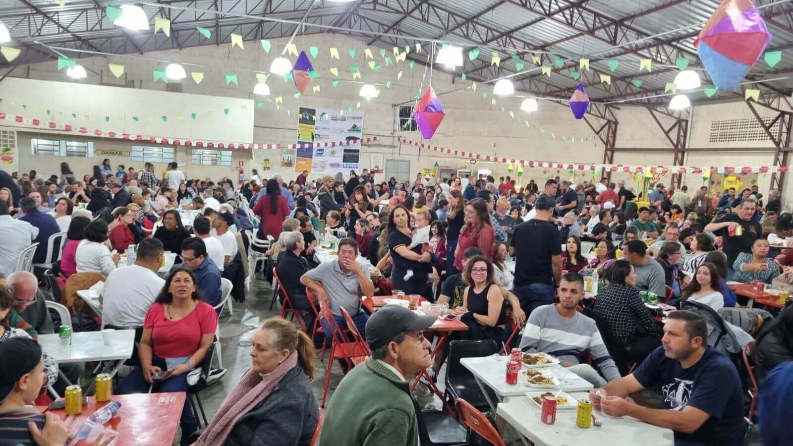 Festa decorada com bandeirinhas no teto, típicos de festas juninas ou comemorações religiosas. Muitas pessoas estão sentadas em mesas em paróquia de Jundiaí.