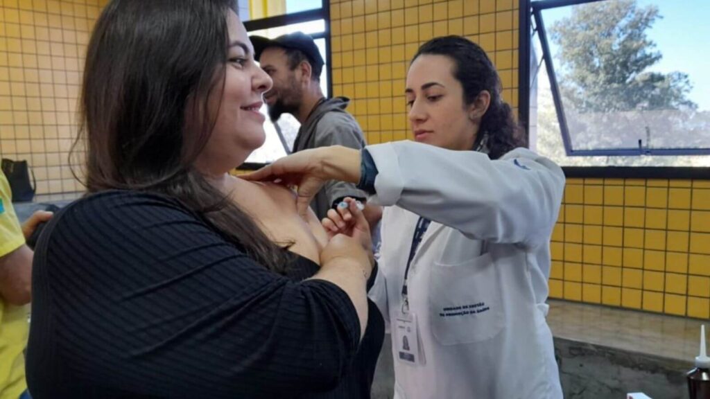 Profissional de saúde vestida com um jaleco branco com o logotipo do SUS aplicando uma vacina da gripe em uma mulher de blusa preta.