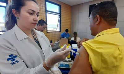 Profissional de saúde, vestida com um jaleco branco com o logotipo do SUS, administrando uma vacina a um homem de camiseta amarela, funcionário do transporte público de Jundiaí.
