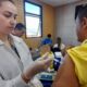 Profissional de saúde, vestida com um jaleco branco com o logotipo do SUS, administrando uma vacina a um homem de camiseta amarela, funcionário do transporte público de Jundiaí.