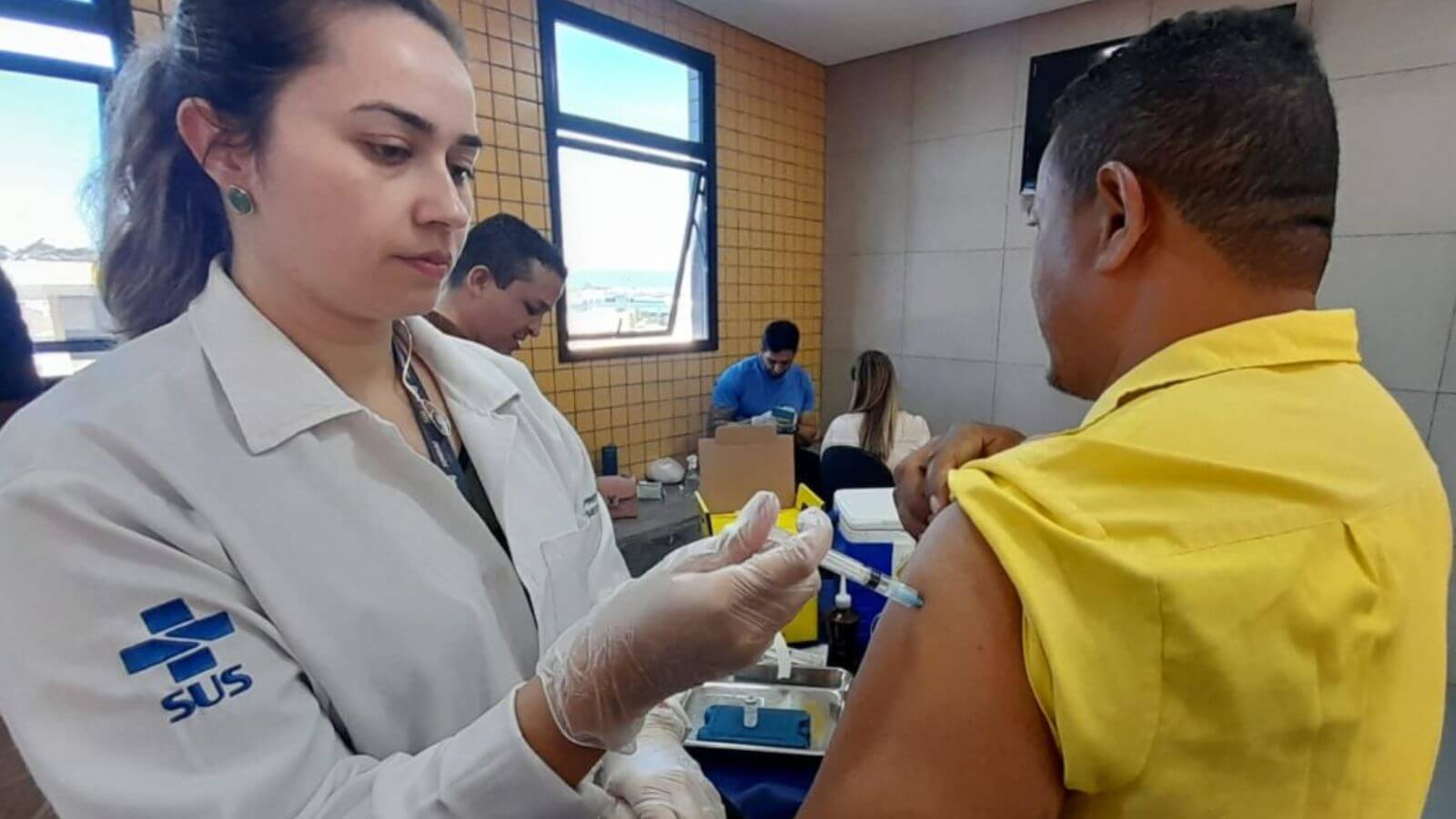 Profissional de saúde, vestida com um jaleco branco com o logotipo do SUS, administrando uma vacina a um homem de camiseta amarela, funcionário do transporte público de Jundiaí.