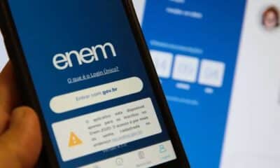 Uma mão segura um smartphone exibindo a tela de login das inscrições do ENEM 2024, com um fundo desfocado mostrando uma interface semelhante em uma tela maior. Enem 2024.