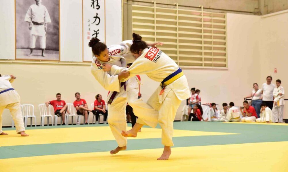 Duas mulheres em um combate durante uma competição de judô, usando quimonos brancos com detalhes em vermelho do Sesi-SP. Elas estão em um tatame de cor amarela e verde, enquanto outros competidores e espectadores assistem ao fundo.