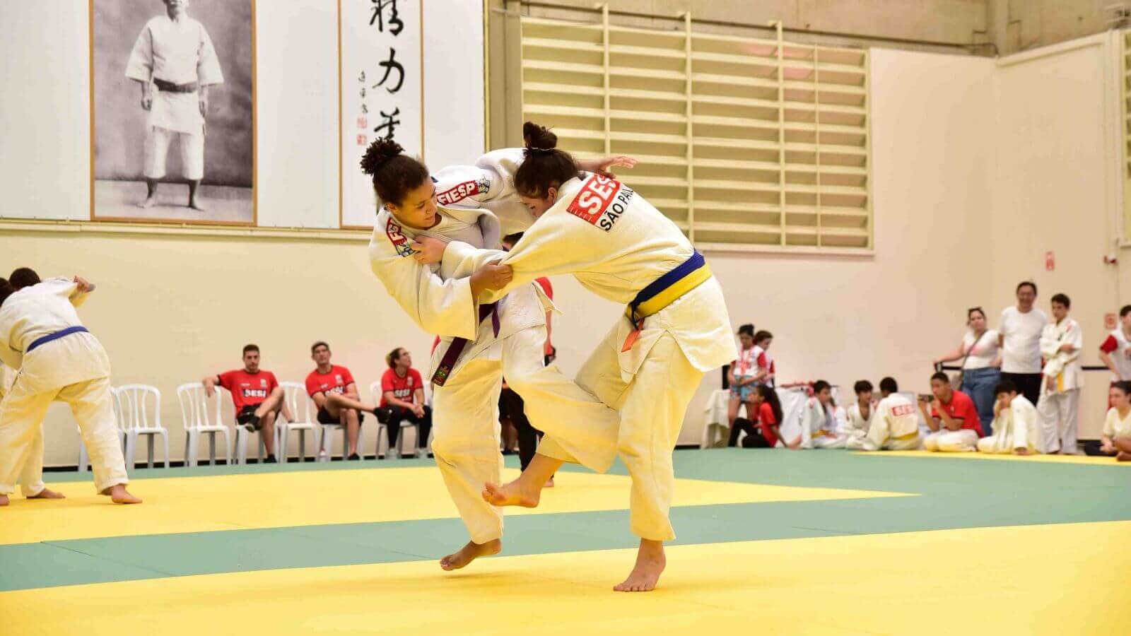 Duas mulheres em um combate durante uma competição de judô, usando quimonos brancos com detalhes em vermelho do Sesi-SP. Elas estão em um tatame de cor amarela e verde, enquanto outros competidores e espectadores assistem ao fundo.