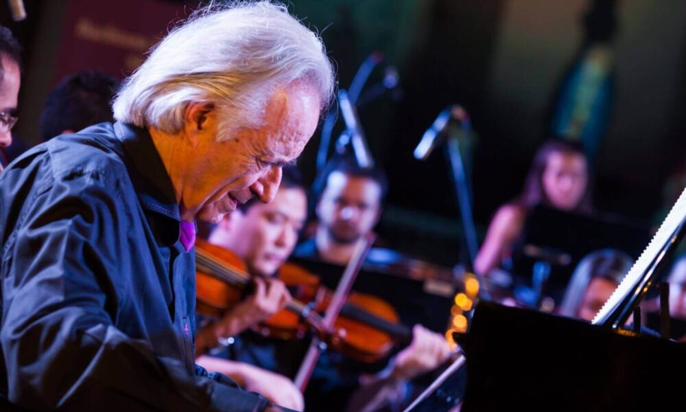 Maestro João Carlos Martins, se apresentando em um concerto, tocando piano. Ao fundo, membros da Bachiana Filarmônica estão tocando instrumentos de corda, como violinos.