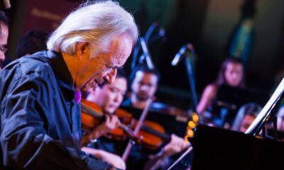 Maestro João Carlos Martins, se apresentando em um concerto, tocando piano. Ao fundo, membros da Bachiana Filarmônica estão tocando instrumentos de corda, como violinos.