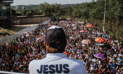 Líder de costas com camiseta "Jesus Vencedor" discursando para multidão na Marcha para Jesus, com pessoas e guarda-chuvas coloridos.