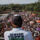 Líder de costas com camiseta "Jesus Vencedor" discursando para multidão na Marcha para Jesus, com pessoas e guarda-chuvas coloridos.