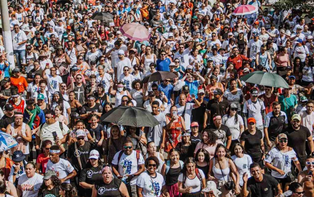 Multidão participando da Marcha para Jesus em um dia ensolarado, com muitas pessoas usando camisetas temáticas e carregando guarda-chuvas.