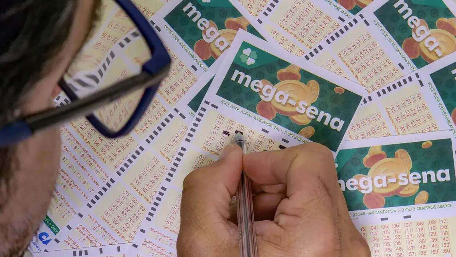 Homem marca números em bilhetes da Mega-Sena espalhados em mesa.