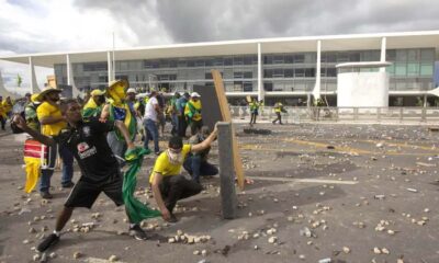 Manifestantes em frente à Praça dos Três Poderes, em Brasília, com alguns participantes jogando objetos e outros se protegendo com barricadas improvisadas nos atos golpistas de 8 de janeiro.