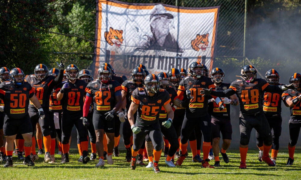 Equipe do Ocelots Futebol Americano entrando em campo com uniformes pretos e laranja antes do jogo, com bandeira do time ao fundo