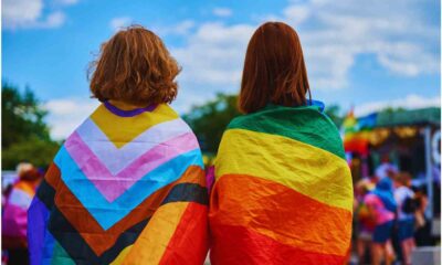Duas pessoas de costas usando bandeiras do orgulho LGBTQIA+ na Parada LGBT+ 2024, uma com a bandeira do orgulho progressista e outra com a bandeira do arco-íris.