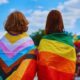 Duas pessoas de costas usando bandeiras do orgulho LGBTQIA+ na Parada LGBT+ 2024, uma com a bandeira do orgulho progressista e outra com a bandeira do arco-íris.