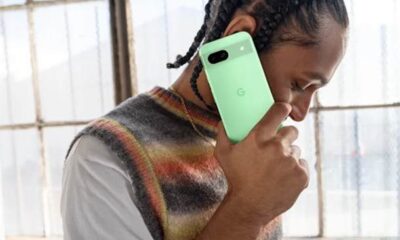 Homem segurando um smartphone Google Pixel 8 verde, encostado ao ouvido, em um ambiente interno iluminado, com cabelo trançado e usando um colete listrado.
