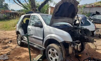 Veículo prateado parcialmente desmontado em um terreno aberto de desmanche clandestino em Campo Limpo Paulista. O capô do carro está levantado, expondo o motor, e as portas estão removidas.