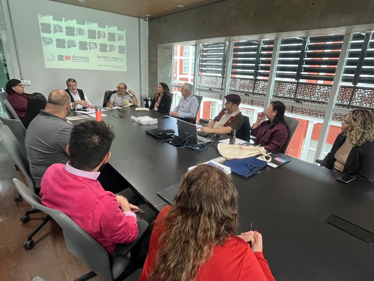 Reunião no Escritório de Projetos da Região Metropolitana de Jundiaí, com 11 pessoas sentadas ao redor de uma mesa grande, discutindo sobre projetos regionais de São Paulo.