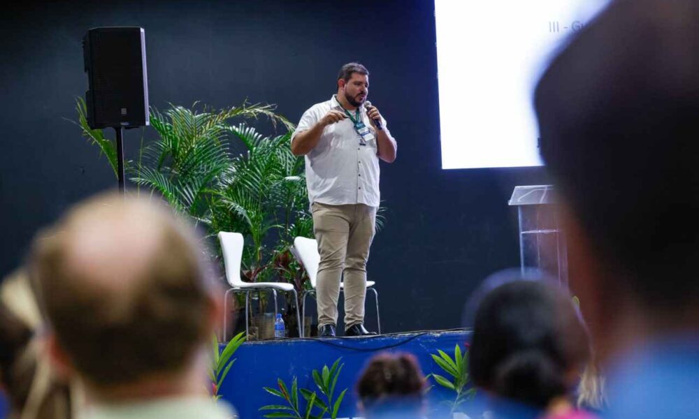 Homem apresentando no Fórum Paulista de Arborização Urbana, com plateia ao fundo, vegetação ao redor e tela de projeção.