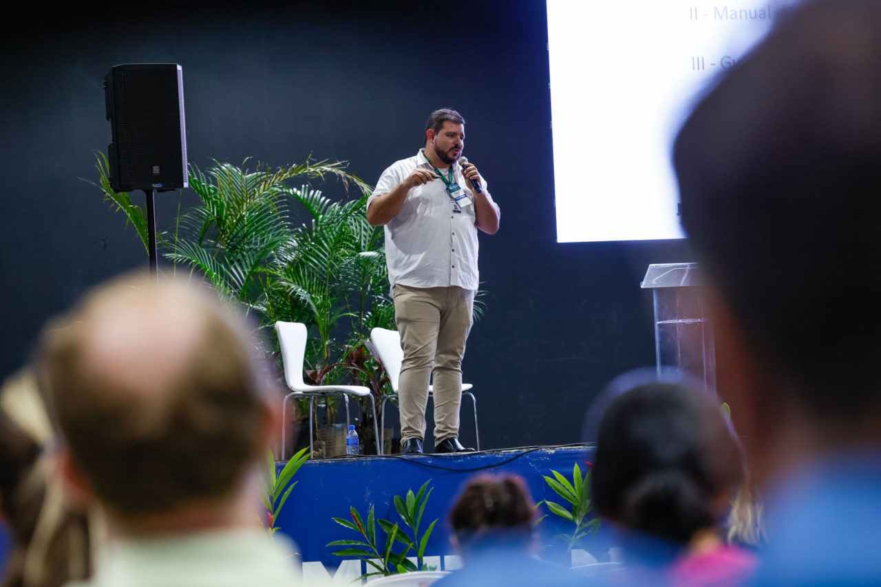 Homem apresentando no Fórum Paulista de Arborização Urbana, com plateia ao fundo, vegetação ao redor e tela de projeção.