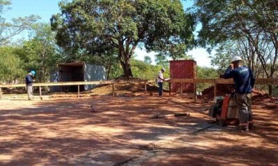 Obras da Cidade das Crianças em Jundiaí, mostrando trabalhadores em um canteiro de obras cercado por árvores, com contêineres ao fundo.