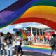 Grupo de pessoas segurando uma grande bandeira de arco-íris, que é símbolo do movimento LGBTQIA+.