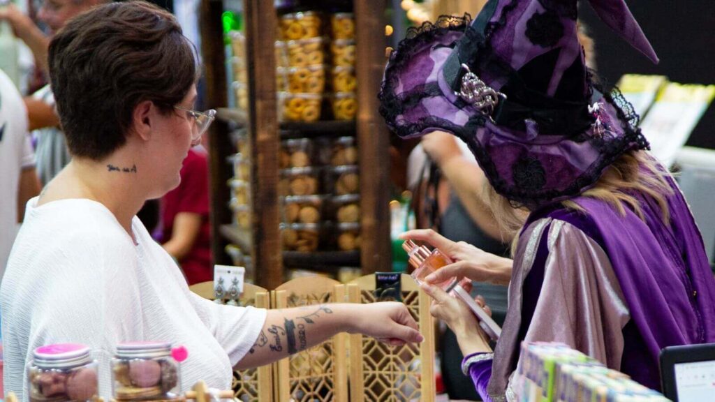 Feira Villa Mistika em Jundiaí, com expositora fantasiada de bruxa aplicando produto no braço de visitante.