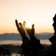 Pai de Santo em silhueta, segurando o sol com as mãos durante o nascer do sol, com montanhas ao fundo.