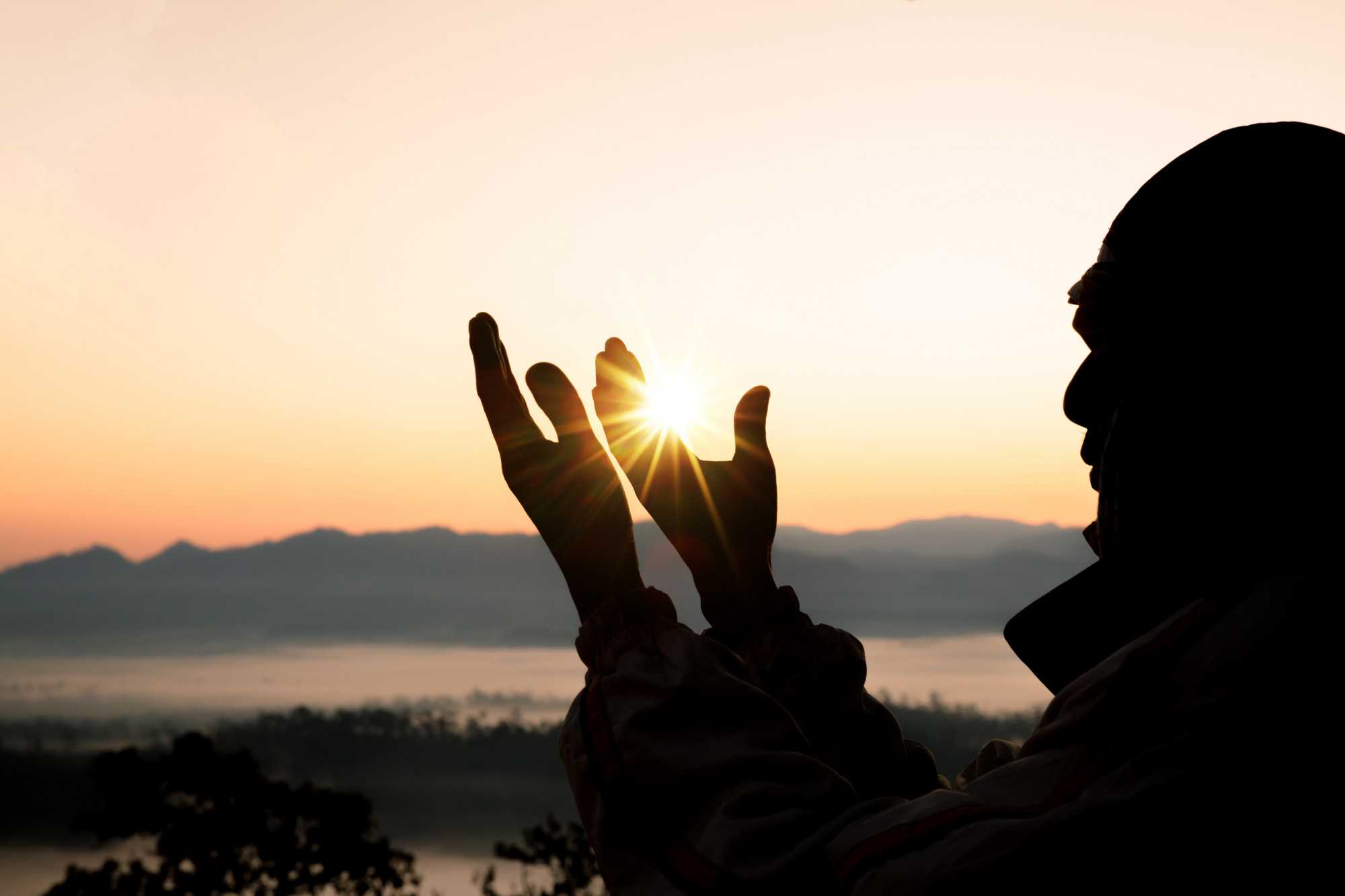 Pai de Santo em silhueta, segurando o sol com as mãos durante o nascer do sol, com montanhas ao fundo.