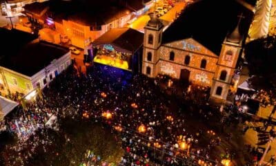 Imagem aérea noturna do Festival de Inverno de Cabreúva 2024, com palco iluminado, multidão e igreja histórica ao fundo.