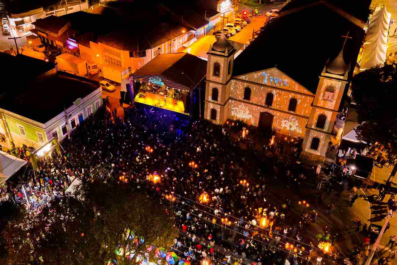 Imagem aérea noturna do Festival de Inverno de Cabreúva 2024, com palco iluminado, multidão e igreja histórica ao fundo.