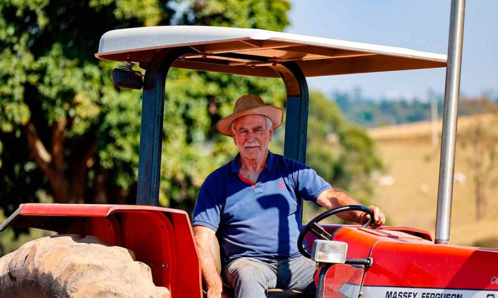 Homem idoso usando chapéu dirige um trator vermelho Massey Ferguson em uma fazenda, com árvores ao fundo em um dia ensolarado, representando os agricultores de Jundiaí.