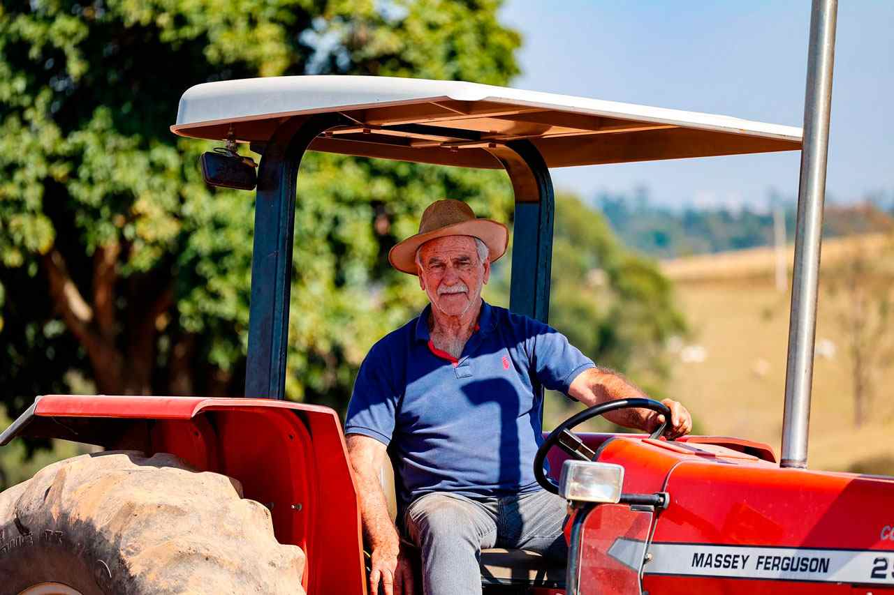 Homem idoso usando chapéu dirige um trator vermelho Massey Ferguson em uma fazenda, com árvores ao fundo em um dia ensolarado, representando os agricultores de Jundiaí.