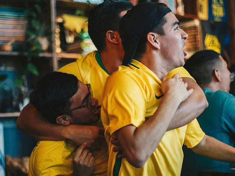 Três torcedores brasileiros comemorando em um bar enquanto assistem a um jogo, representando a emoção das apostas online.