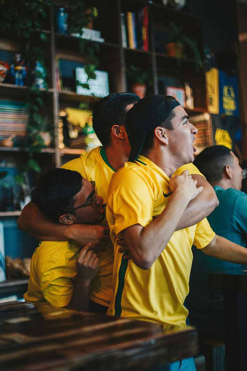 Três torcedores brasileiros comemorando em um bar enquanto assistem a um jogo, representando a emoção das apostas online.