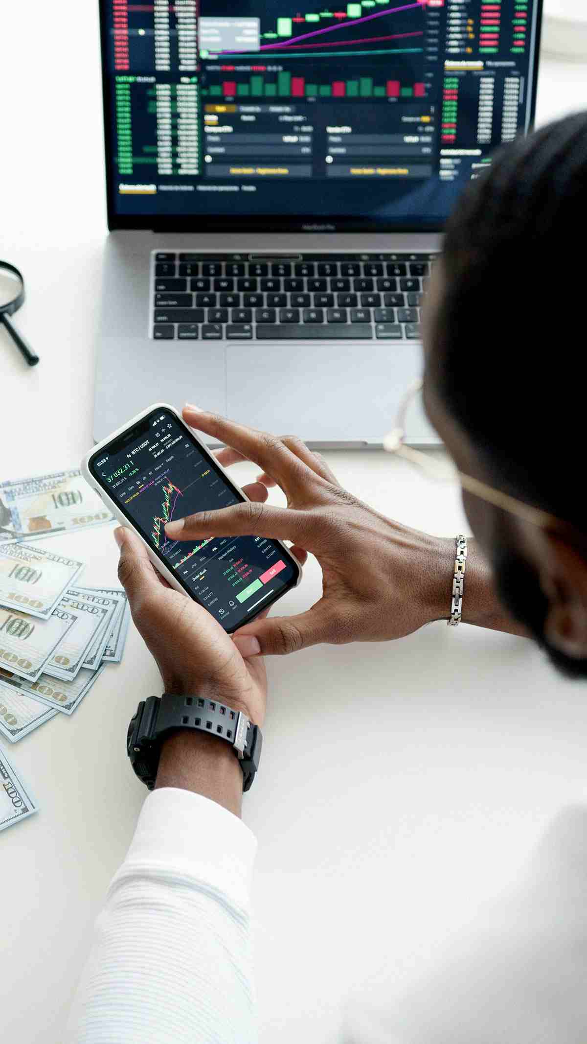 Uma pessoa analisando gráficos de ações ou criptomoedas Bitcoin em um smartphone e laptop, com notas de dólar na mesa.